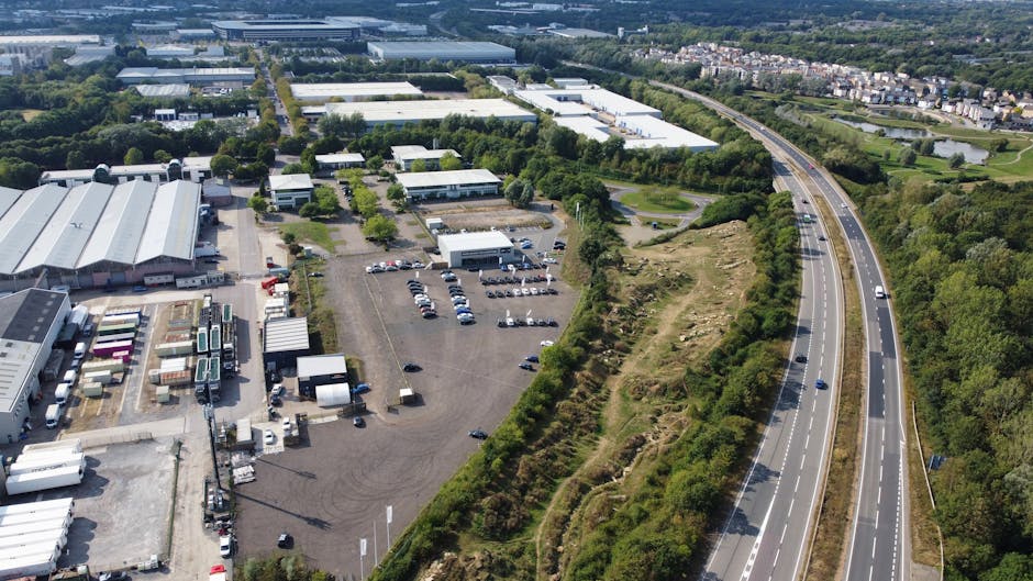Aerial view of a commercial or industrial area situated adjacent to a dual carriageway on the outskirts of Falconwood, Bexley. The image captures multiple large warehouse-style buildings with white and grey exteriors, surrounded by paved loading zones and parking lots with numerous parked vehicles, including trucks and vans. In the foreground, a stretch of open land covered with grass and shrubs runs parallel to the busy road, which has several moving cars. The scene is well-lit with natural daylight, showing the typical environment encountered during a home relocation or furniture transport process. Such an area is representative of the logistics involved in local removals, with vehicles arriving and departing, and space available for loading, unloading, and organizing household possessions, which Man with van Falconwood manages as part of their property moving and packing services.