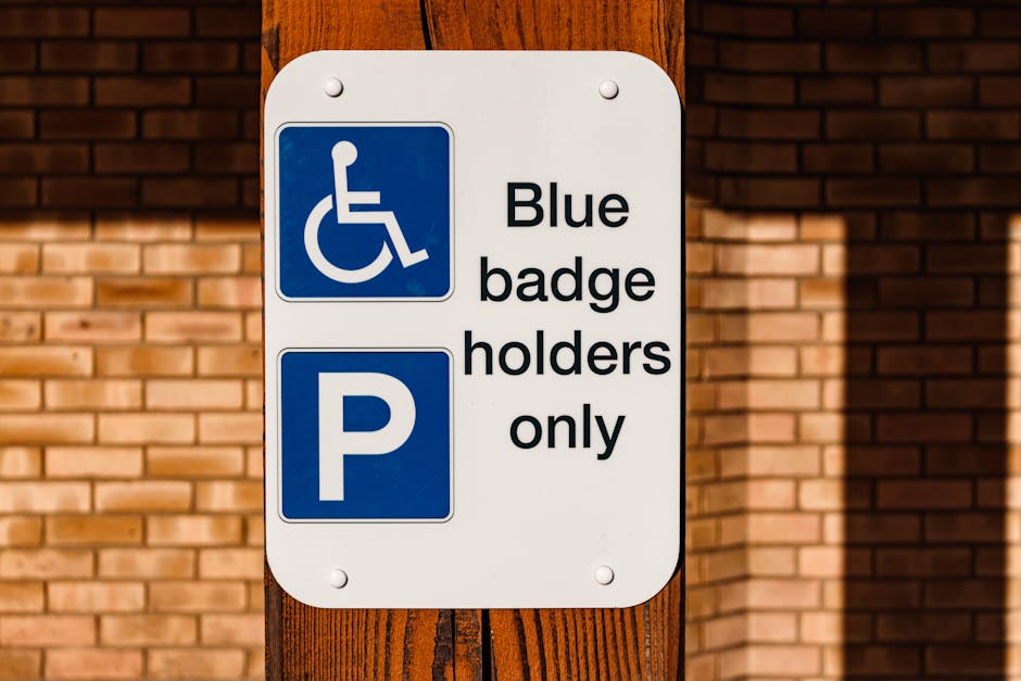 A close-up image of a white sign mounted on a wooden post with bolts, positioned in front of a red brick wall. The sign features a blue wheelchair accessibility symbol at the top left, indicating designated parking for disabled badge holders, and a blue parking symbol with a white 'P' below it. The sign contains the text 'Blue badge holders only' in black lettering. The environment appears to be outdoors with sunlight casting shadows on the wall and sign. This signage is relevant to vehicle parking regulations, which can be part of the logistics involved in home relocation or moving services provided by Man with van Falconwood, ensuring accessible parking arrangements during furniture transport and packing and moving operations.