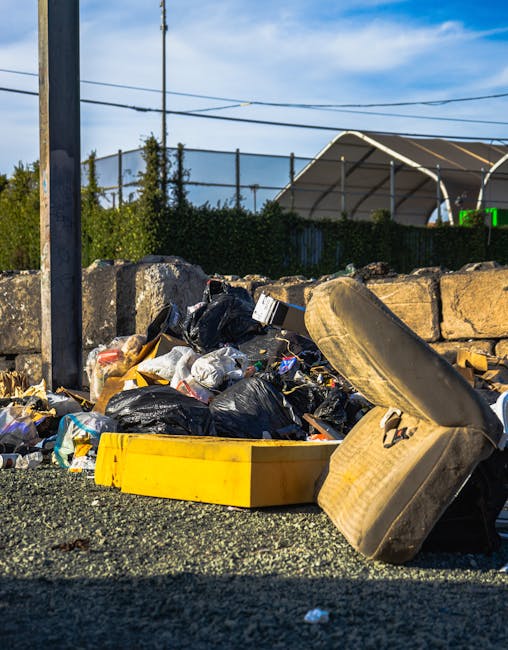 A row of large white and dark grey wheelie bins with closed lids are positioned next to a dark metal storage container on a concrete pavement, with some of the bins labelled for waste disposal such as