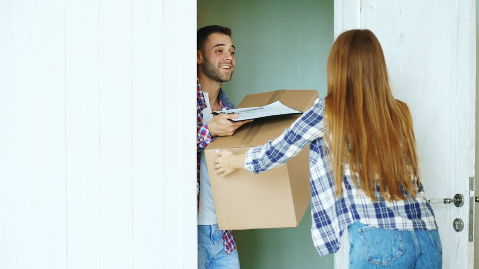 A man wearing a yellow T-shirt and blue jeans is standing inside a room during a home relocation process, holding a clipboard and a black marker for taking notes or marking inventory. Behind him, ther