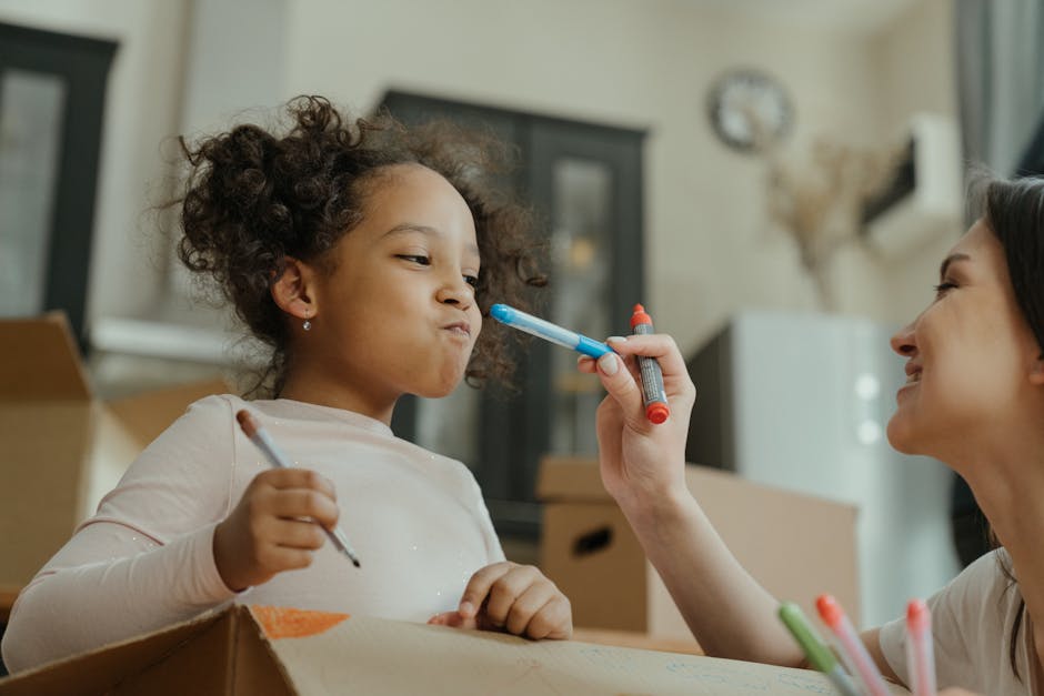 Image showing a young girl with curly dark hair and light skin inside a home during a packing and moving process. She is holding a pen and standing next to a cardboard box. An adult woman with light skin and dark hair, smiling and holding a blue marker, is gently touching the girl's face with it. The background includes a living room with a dark cabinet, a white appliance, a wall clock, and a decorative wall hanging. The scene is well-lit with natural light coming through a window covered with light curtains. The setting reflects a household preparing for a home relocation, with packing materials and furniture visible, captured during an informal packing or decorating activity, illustrating the logistics of furniture transport and moving preparations by Man with van Falconwood.