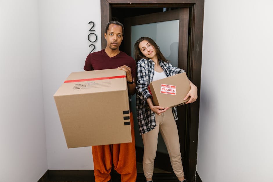 Two movers from R.C. Moving Ltd, wearing black caps and T-shirts with the company logo, are outdoors in a bright, modern residential area during daylight. They are carrying cardboard boxes and a large, flat-packed piece of furniture on a trolley, preparing for a home relocation. The boxes are made of brown cardboard, some open and others sealed, with packing tape visible. The trolley has two handles and wheels, assisting in transporting items. They are near a large, wall-mounted mailbox system with multiple small metallic compartments, positioned beside the entrance to a building with white walls and large windows. The background features additional residential buildings with multiple floors and balconies, indicating an urban or suburban environment. The scene captures the unloading or loading process, highlighting efficient furniture transport and packing procedures typical of professional removals services, as provided by Man with Van Falconwood, supporting efficient short-notice moves and home relocations.