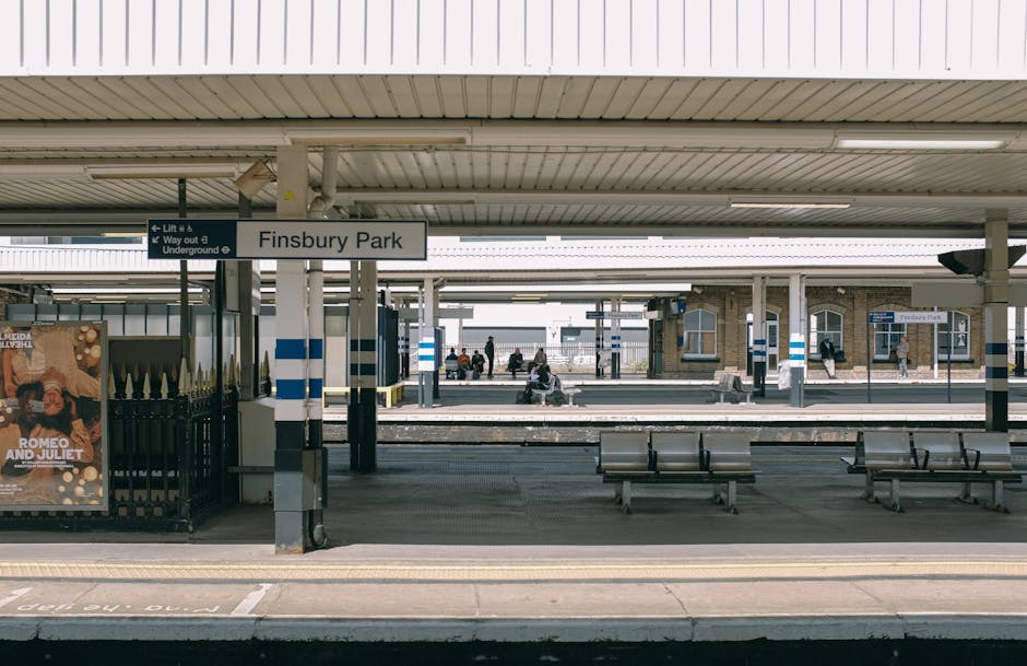 View of Finsbury Park train station platform with several empty metal benches positioned along the concrete surface, covered by a large corrugated metal canopy supported by metal pillars painted with blue and white stripes. A white sign with black text indicates the station name as 'Finsbury Park' and provides directions to lift and underground access. The platforms are separated by railway tracks, with some passengers sitting on benches and others walking along the platform. In the foreground, there are a few cardboard boxes and protective blankets, possibly prepared for a home relocation or furniture transport, positioned near the edge of the platform. The environment appears clean and well-lit with natural daylight, indicating daytime hours. Occasionally, companies like Man with Van Falconwood conduct such transport activities, including packing, loading, and moving furniture during property relocations, similar to the context suggested by the 'FALCONWOOD' local moving guide page.