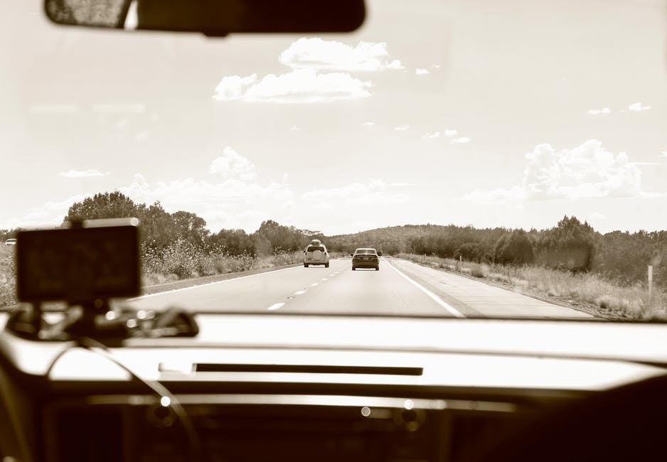 A view from inside a vehicle on a clear day showing a two-lane road ahead with two cars driving in the same direction, surrounded by trees and low bushes on both sides. The dashboard of the vehicle includes a mounted GPS device and some cables, indicating the driver may be using navigation technology. The sky is partly cloudy with large, fluffy clouds scattered across it, and bright sunlight illuminates the scene. This image captures a moment in a home relocation journey, with the focus on the travel environment during furniture transport or moving logistics, highlighting the road and vehicle interior within the context of house removals and packing and moving activities. The setting suggests an ongoing journey to or from a house, emphasizing the importance of professional removals and transportation services such as those offered by Man with van Falconwood.
