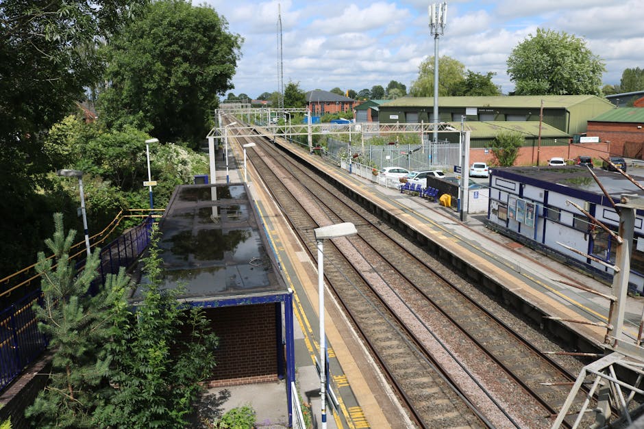 View of Finsbury Park train station platform with several empty metal benches positioned along the concrete surface, covered by a large corrugated metal canopy supported by metal pillars painted with blue and white stripes. A white sign with black text indicates the station name as 'Finsbury Park' and provides directions to lift and underground access. The platforms are separated by railway tracks, with some passengers sitting on benches and others walking along the platform. In the foreground, there are a few cardboard boxes and protective blankets, possibly prepared for a home relocation or furniture transport, positioned near the edge of the platform. The environment appears clean and well-lit with natural daylight, indicating daytime hours. Occasionally, companies like Man with Van Falconwood conduct such transport activities, including packing, loading, and moving furniture during property relocations, similar to the context suggested by the 'FALCONWOOD' local moving guide page.