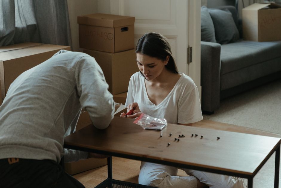 A woman with long dark hair, wearing a white t-shirt, sits on the floor of a living room surrounded by packed cardboard boxes and furniture, as she sorts small screws or hardware from a clear plastic bag onto a wooden table. A man, dressed in a light-colored hoodie, is partially visible, leaning over the table and assisting with packing or assembling items. Behind them, a grey sofa with cushions, a window with curtains, and additional moving boxes are visible, indicating an ongoing moving process. The scene captures the interior environment where home relocation preparations are taking place, with focus on packing and handling furniture during the loading process, which is typical of moving services provided by Man with van Falconwood.