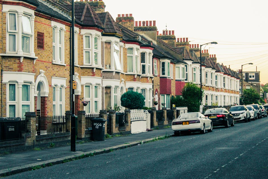 A row of terraced residential houses with brick facades and white bay windows facing a street, lined with parked cars on both sides, including sedans and hatchbacks. The sidewalk features small front gardens with decorative fences, some with potted plants and shrubbery. A black wheelie bin is visible near a lamppost, and the street is illuminated by streetlights and natural evening light. The scene depicts a typical neighbourhood prepared for a moving or home relocation process, with clear access to the front doors and the pavement for loading and unloading furniture or boxes. This setting reflects the environment where professional removals, such as those provided by Man with van Falconwood, would facilitate house moves involving packing, furniture transport, and parking considerations on Welling Road, SE9.