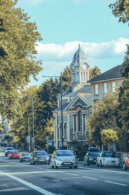 A row of terraced residential houses with brick facades and white bay windows facing a street, lined with parked cars on both sides, including sedans and hatchbacks. The sidewalk features small front gardens with decorative fences, some with potted plants and shrubbery. A black wheelie bin is visible near a lamppost, and the street is illuminated by streetlights and natural evening light. The scene depicts a typical neighbourhood prepared for a moving or home relocation process, with clear access to the front doors and the pavement for loading and unloading furniture or boxes. This setting reflects the environment where professional removals, such as those provided by Man with van Falconwood, would facilitate house moves involving packing, furniture transport, and parking considerations on Welling Road, SE9.