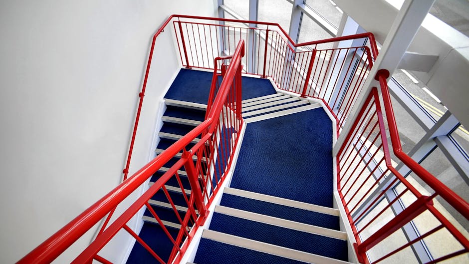 Aerial view of a multi-story internal staircase in a residential or commercial building, featuring red metal handrails and white framing. The staircase is covered with blue carpeted treads, and the surrounding walls are painted white. Natural light from large floor-to-ceiling windows illuminates the staircase area, which appears to be part of a house or apartment building undergoing a furniture transport or home relocation process. The image captures the top landing and the descending staircase, with no objects or furniture visible, focusing solely on the structural elements and safety railings. Occasionally, [COMPANY_NAME], such as Man with van Falconwood, may use such staircases during the loading and unloading phases of moving services, especially when navigating tight access points in renovations or relocations.