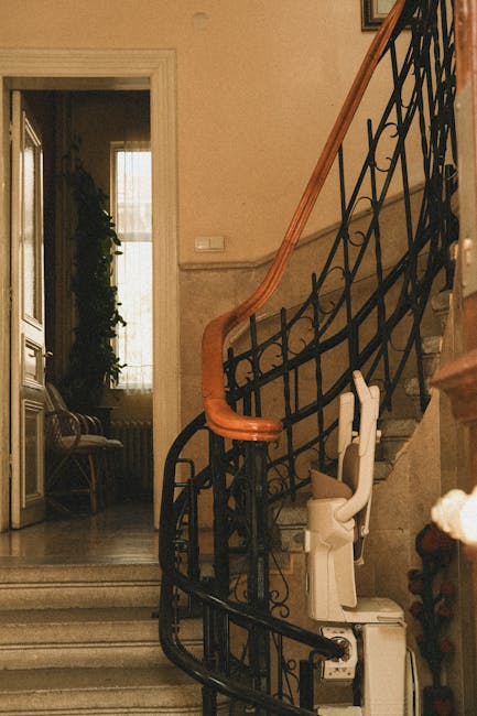 A spiral staircase with a wooden handrail and ornate black metal balustrades ascends inside a residential property, with a white stairlift attached to the side of the stairs. Behind the staircase, a doorway opens into a room with a window covered by sheer curtains, allowing natural light to illuminate the space. Near the base of the stairs, a commercial moving box made of cardboard is positioned on the floor, suggesting packing or ongoing furniture transport activities as part of a home relocation process. The setting appears clean and well-maintained, with neutral interior walls, and the environment indicates a typical indoor scene during a household move. This image relates to house removals and furniture transport, emphasizing the logistics of packing and moving within a residential setting, as managed by Man with van Falconwood in their specialist removals services.