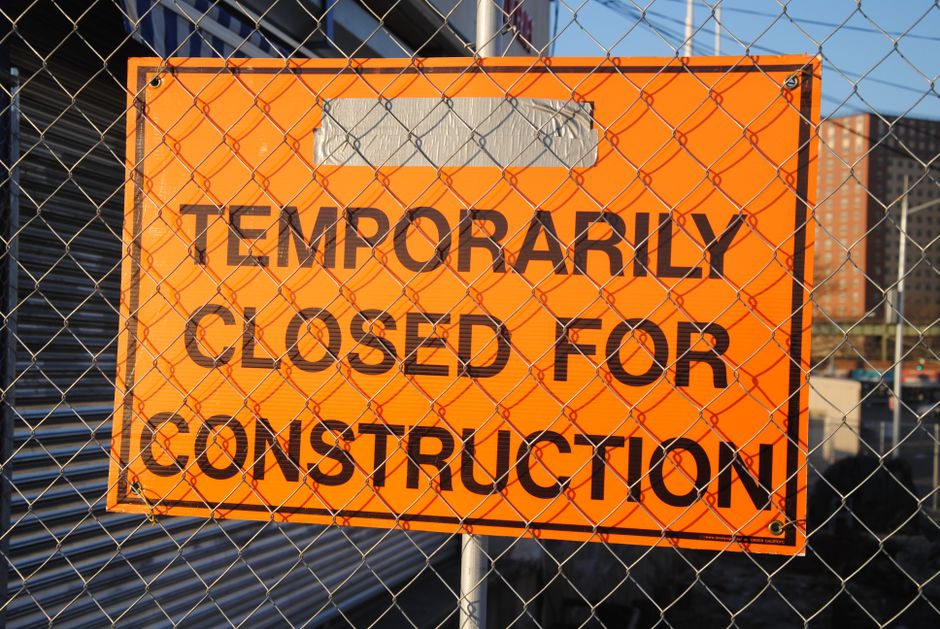 A rectangular orange sign with bold black text reading 'Temporarily Closed for Construction' is attached to a metal fence with a diamond-shaped wire mesh pattern. The sign is positioned in front of a construction site, which is partially visible through the fence. In the background, there are high-rise buildings, construction materials, and machinery, indicating ongoing building work. The scene is outdoors with natural daylight illuminating the sign and the surrounding area. The placement of the sign suggests that the location is temporarily inaccessible, consistent with house removals and logistics support from [COMPANY_NAME], which manages property relocations and transportation of furniture and boxes. The fencing and signage indicate a controlled and secure environment typical of relocation projects requiring careful planning of access routes and permits, relevant to the page about 'Temporary Road Closure and Moving Permits in SE9, Falconwood'.