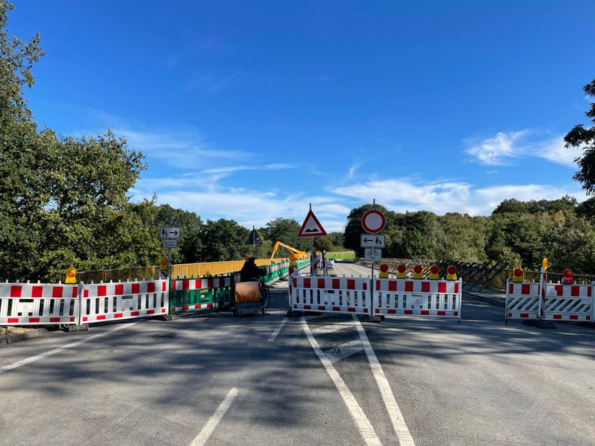 Photograph of a street undergoing temporary road closure for house removals and moving logistics in SE9, Falconwood. The road is blocked by white and red plastic barriers and green metal fencing, with several signs indicating no entry and warning of construction activity. A wheeled trolley loaded with cardboard boxes and wrapped furniture is positioned near the barriers, suggesting ongoing packing or furniture transport. In the background, a yellow crane is visible, likely assisting with the lifting and movement of large household items onto a waiting van or truck. The scene is outdoors, surrounded by trees and under a bright blue sky with scattered clouds, illustrating an organized effort by Man with van Falconwood to facilitate home relocation and furniture transport services, while managing traffic and safety during the process.