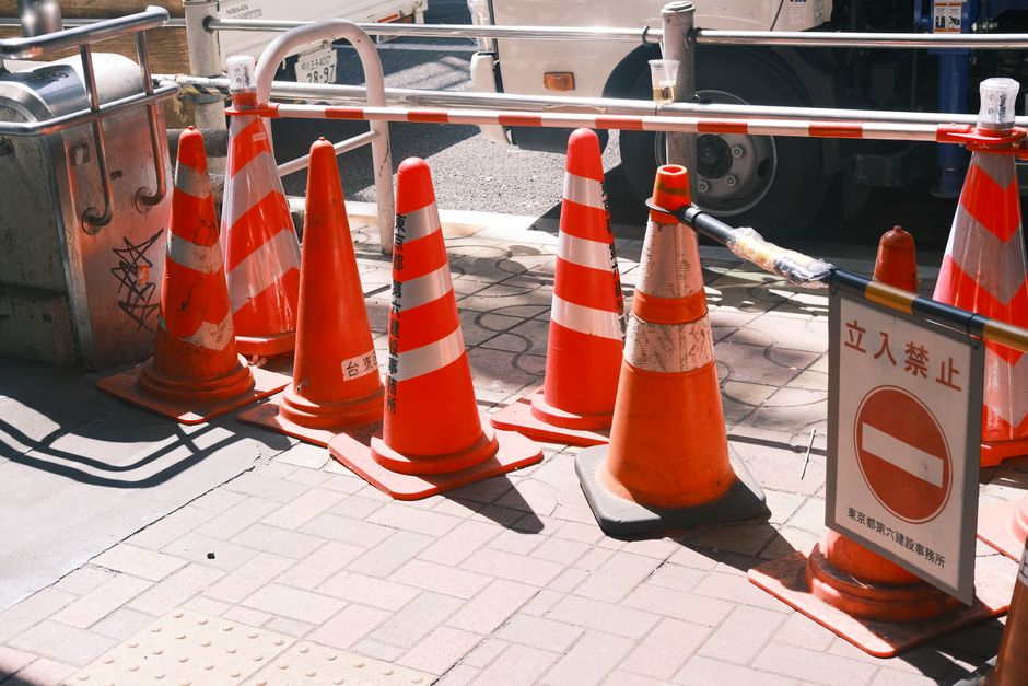 A rectangular orange sign with bold black text reading 'Temporarily Closed for Construction' is attached to a metal fence with a diamond-shaped wire mesh pattern. The sign is positioned in front of a construction site, which is partially visible through the fence. In the background, there are high-rise buildings, construction materials, and machinery, indicating ongoing building work. The scene is outdoors with natural daylight illuminating the sign and the surrounding area. The placement of the sign suggests that the location is temporarily inaccessible, consistent with house removals and logistics support from [COMPANY_NAME], which manages property relocations and transportation of furniture and boxes. The fencing and signage indicate a controlled and secure environment typical of relocation projects requiring careful planning of access routes and permits, relevant to the page about 'Temporary Road Closure and Moving Permits in SE9, Falconwood'.