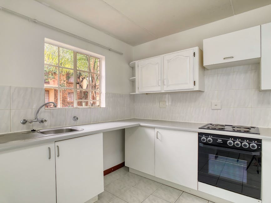 A kitchen interior with white cabinetry and tiled walls viewed from the corner, featuring a stainless steel single sink with a curved faucet beneath a window that allows natural light and offers a view of trees outside. The countertop is clear, with some gaps for appliances, including a black freestanding electric oven with four burners on top. Above the countertop, there are wall-mounted cupboards with paneled doors; one has a slight decorative arch, and another is a simple rectangular design. Inside the room, a white cabinet door is partially visible beneath the countertop, possibly concealing storage or plumbing connections. The kitchen floor is tiled with light grey ceramic tiles matching the wall tiles, and the room's overall appearance is clean, organized, and prepared for home relocation or packing and moving activities, as part of a house removal process by Man with van Falconwood. The lighting appears natural, coming through the window, with no other figures or objects visible.