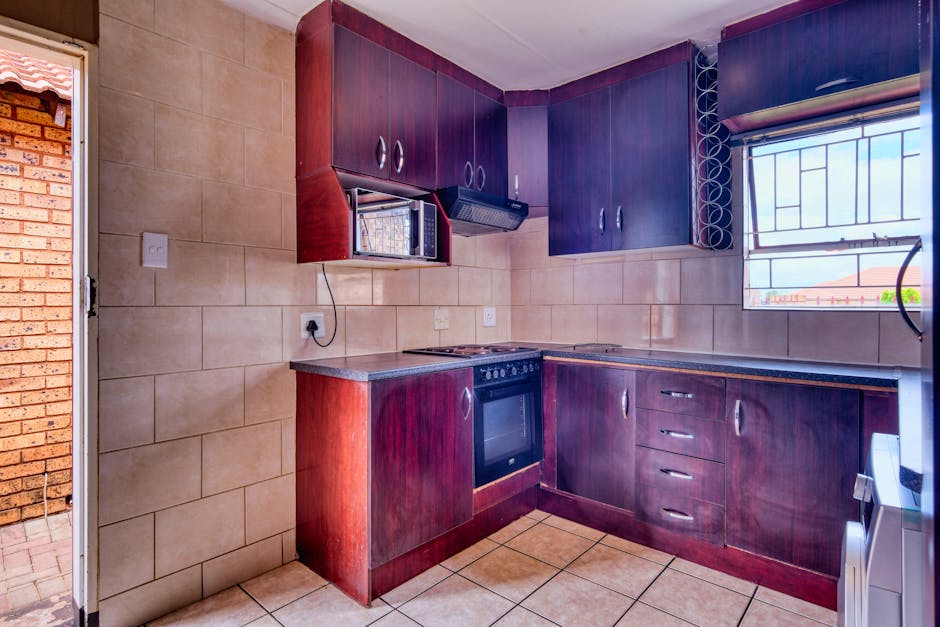A kitchen interior being prepared for a house relocation with dark wooden cabinetry mounted above beige tiled walls and corresponding lower cabinets, including a built-in oven and stovetop. The space features a small microwave oven installed beneath an upper cabinet, with a power cord plugged into an outlet on the wall. To the right, a window with metal security bars allows natural light into the room, revealing a view of neighboring rooftops. The kitchen floor is tiled in a neutral color, and the room appears tidy, ready for packing and moving activities. Outside the open door, part of an outdoor area with exposed brickwork can be seen, suggesting the transition of furniture and boxes from inside to a moving van. This scene is part of a house removal process facilitated by Man with van Falconwood, illustrating a typical workspace during a professional removals and transport operation in Falconwood as part of their urgent same-day move services.